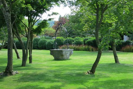 Ein Wasserbecken aus Stein mit einer Verzierung aus Metall auf einem Rasen, umgeben von Bäumen.
