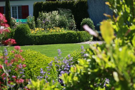 Park oder Garten mit Blumenbeet, weiter Hinten gemähter Rasen und Hecke sowie weiteren Blumen bzw. Rabatten.